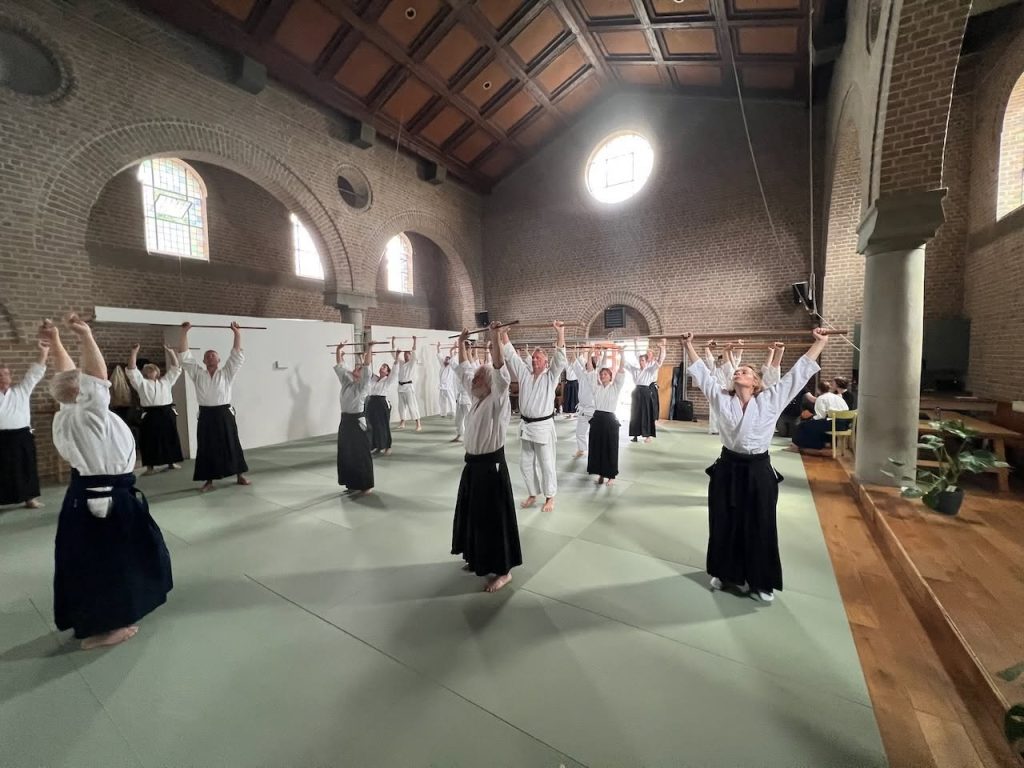 A large group training together with wooden staffs in a bright hall